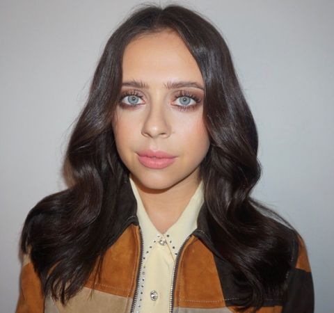 Bel Powley in a brown jacket looks at the camera. 