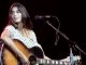 Emmylou Harris in white dress with guitar on hand singing at a stage.