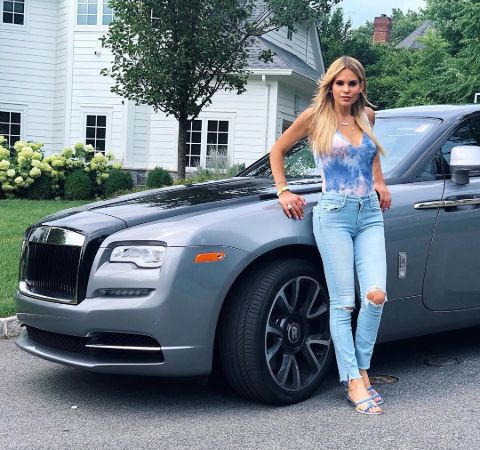 Jackie Goldschneider in a blue top and jeans poses in front of her car.